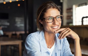 A woman wearing glasses and a light blue shirt smiles while resting her chin on her hand in a cafe setting.